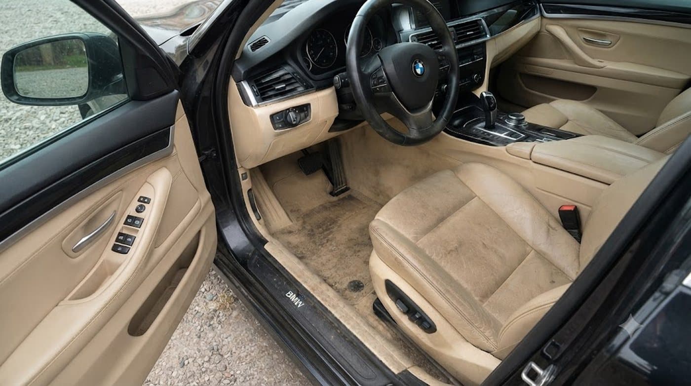 Fahrerseite eines BMW 5er mit beigem Leder: Fußraum und Sitzwange sind großflächig verschmutzt, der Teppich dunkel verfärbt.